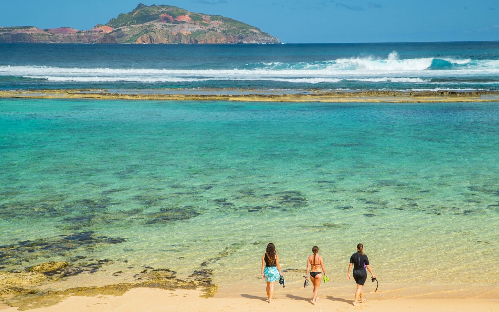 three friends walk towards crystal blue waters on Norfolk Island with views of a neighbouring island in distance
