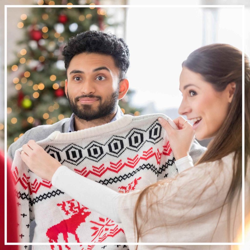 Woman holding up Christmas sweater infront of man who looks uncomfortable about it